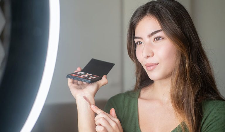 a woman wearing green top holding a blush palette in front of a ring light while vlogging